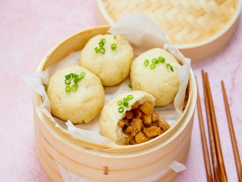 Steamed gluten-free pork buns in bamboo steamer on pink board with chopsticks to the side.