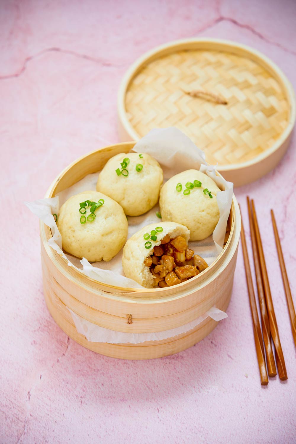 Steamed gluten-free pork buns in bamboo steamer on pink board with chopsticks to the side.