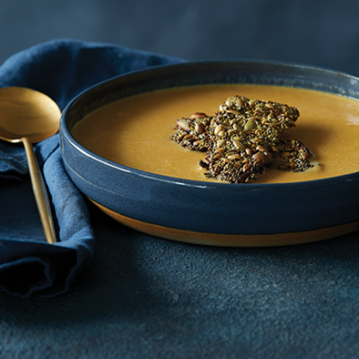 Dark blue bowl of curried sweet potato soup with gluten-free seed crackers and gold spoon.