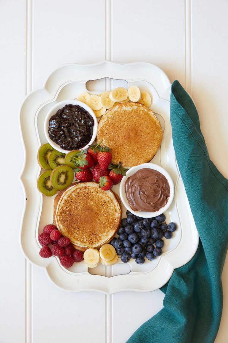 Make ahead gluten-free pancakes on a white platter with assorted fresh fruit, jams and nutella