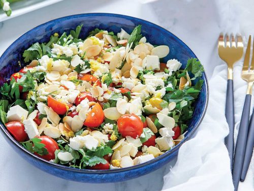 Easy tomato salad topped with feta and flaked almonds in a blue salad bowl with gold cutlery and white napkin.
