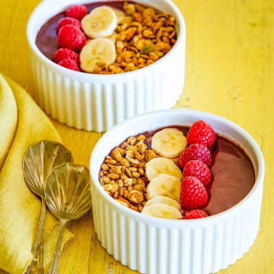 2 x gluten free acai bowls served in white ceramic bowls and topped with fresh raspberries, sliced banana and gluten-free granola. To the side are two vintage spoons and a lemon coloured linen napkin.