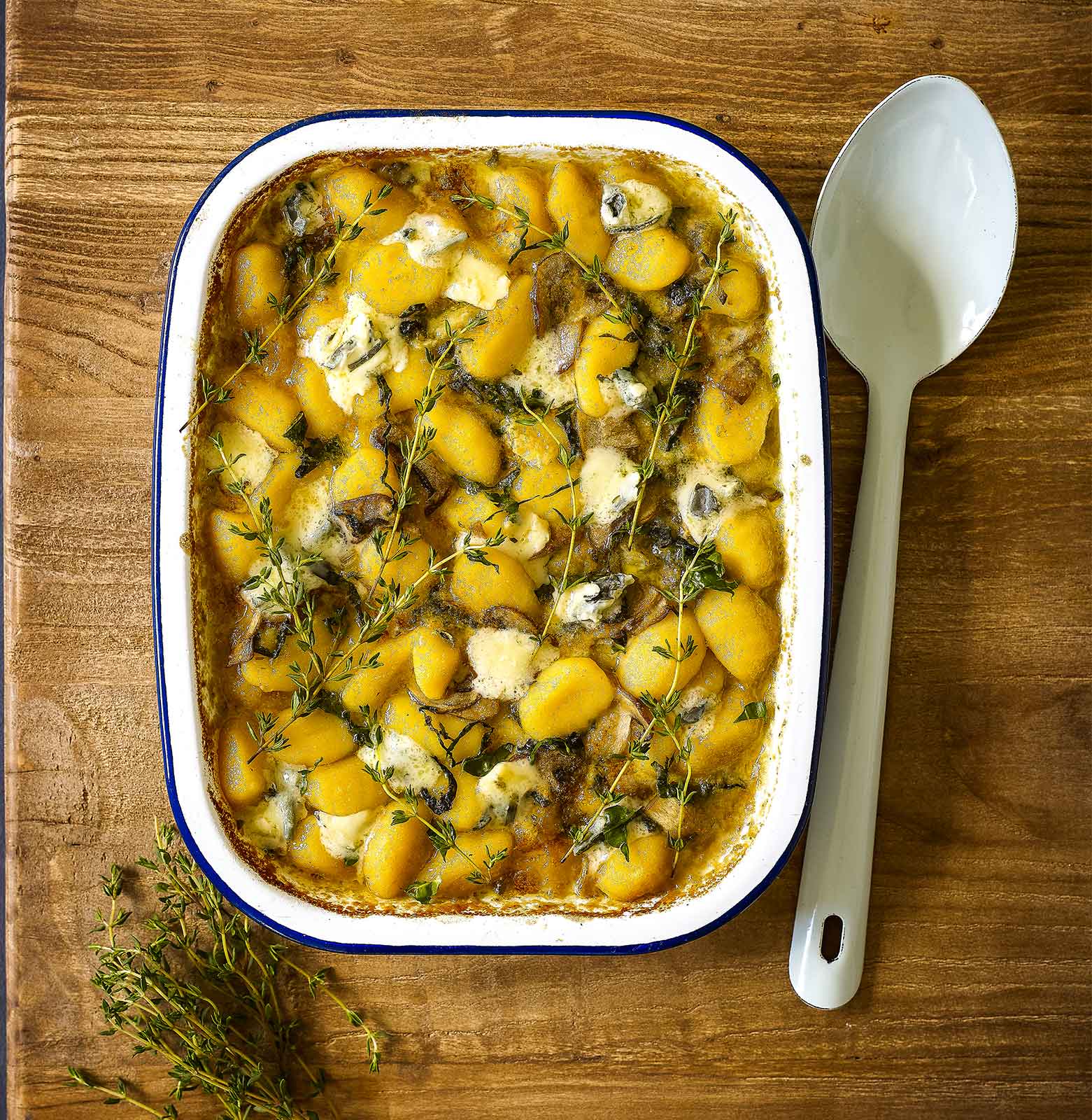 An enamel baking dish filled with gluten-free baked blue cheese gnocchi and topped with lemon thympe. A serving spoon sits to the right ready to serve.