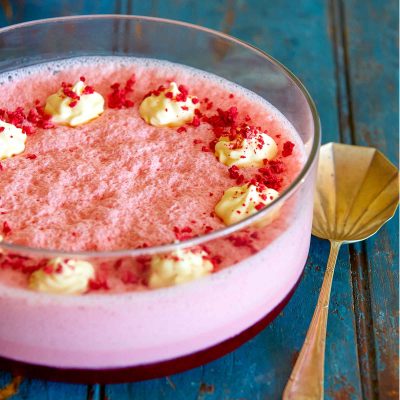 A large glass bowl of raspberry flummery sits on a blue board. The flummery has a bottom layer of raspberry jelly and is topped with the raspberry flummery. Whipped cream is piped on top and it has been sprinkled with crushed freeze dried rasperries.