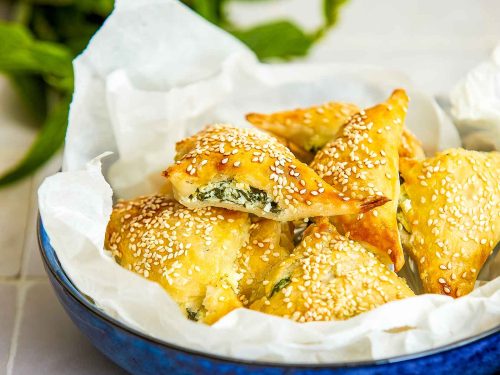 Gluten-free spinach and cheese triangles are piled into a blue bowl lined with paper. A bunch of fresh mint is blurred in the background.