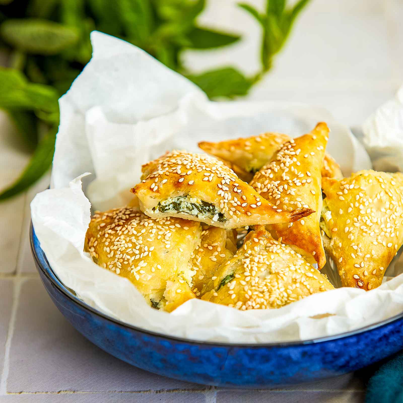 Gluten-free spinach and cheese triangles are piled into a blue bowl lined with paper. A bunch of fresh mint is blurred in the background.