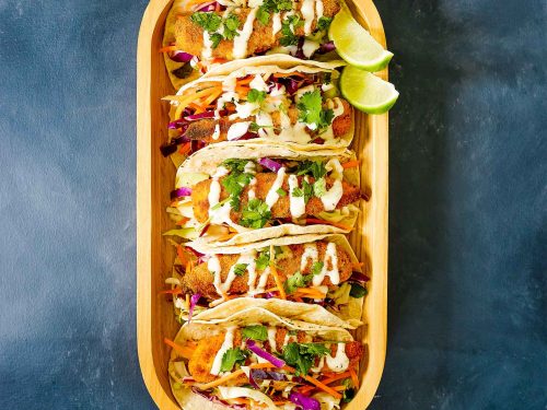 A wooden dish with 5 gluten-free chicken tacos inside. The tacos are drizzled with homemade avocado aioli and have wedges of fresh lime on the side.