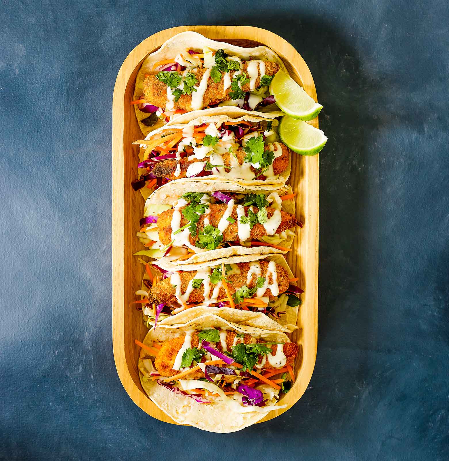 A wooden dish with 5 gluten-free crispy chicken tacos inside. 