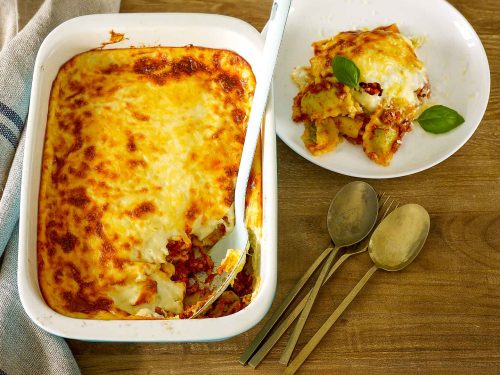 A baking dish with gluten-free spinach and ricotta lasagne rest on a wooden table. A blie enable spoon as removed a scoop which sits on a white plate at the back. Gold spoons are placed to the side of the gluten-free lasagne.