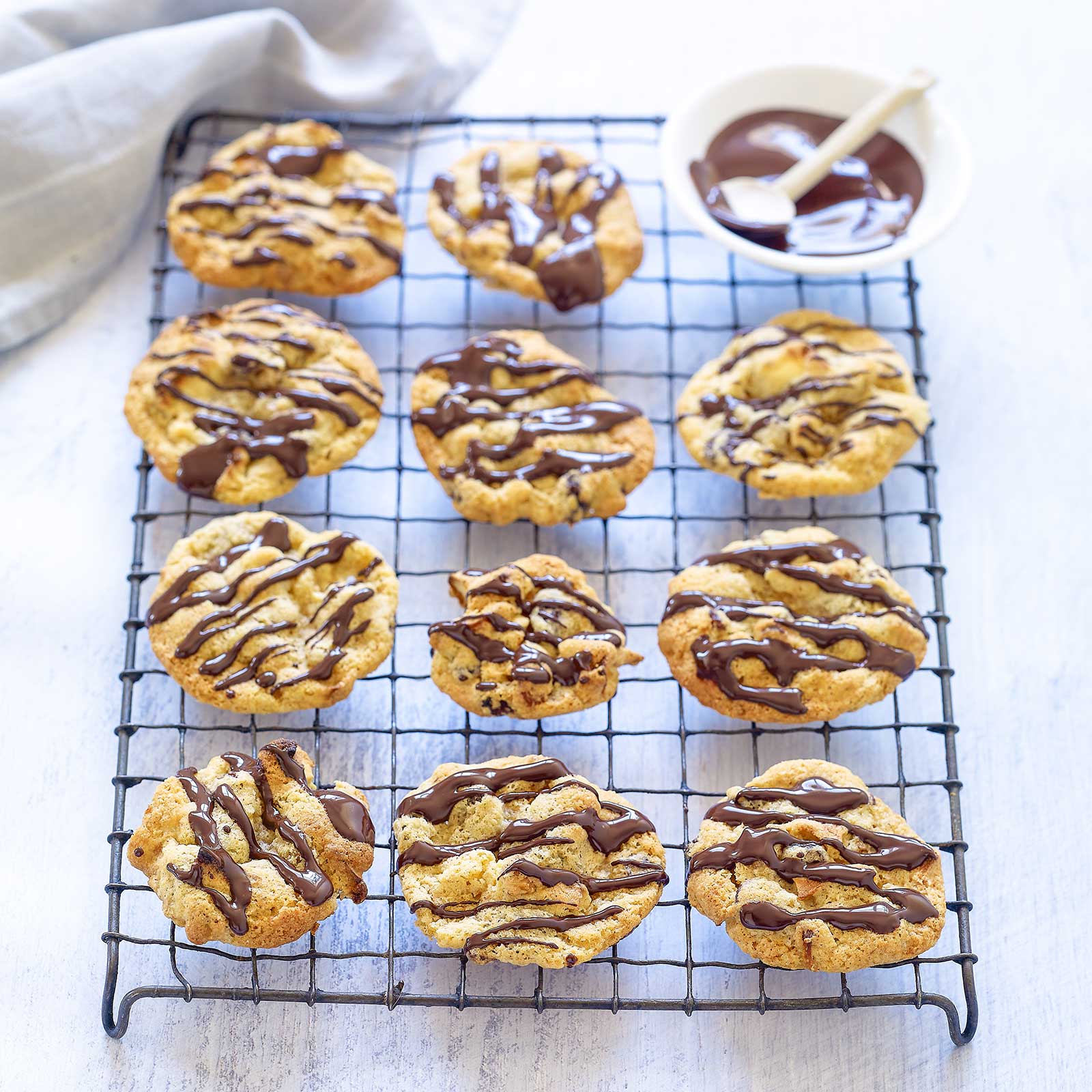 Gluten-free apple and walnut biscuits are arranged on a large wire rack. At the back is a small white bowl with melted chocolate and spoon. The chocolate has been drizzled over the top of the biscuits.