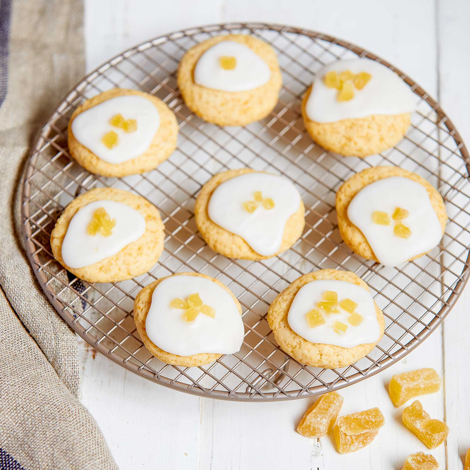 Gluten-free vegan ginger marmalade biscuits are arranged on a round wire cooling tray. A few pieces of crystalised ginger have been sprinkled in front of the tray. The biscuits are round with a white icing and are topped with a few small pieces of crystalised ginger.