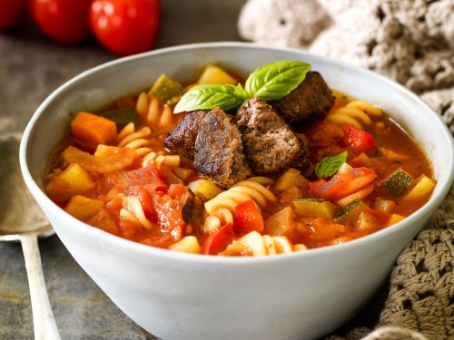 A light blue ceramic bowl is filled with gluten-free minestrone and topped with chopped meatballs and fresh basil leaves. Truss tomatoes sit behind the bowl.