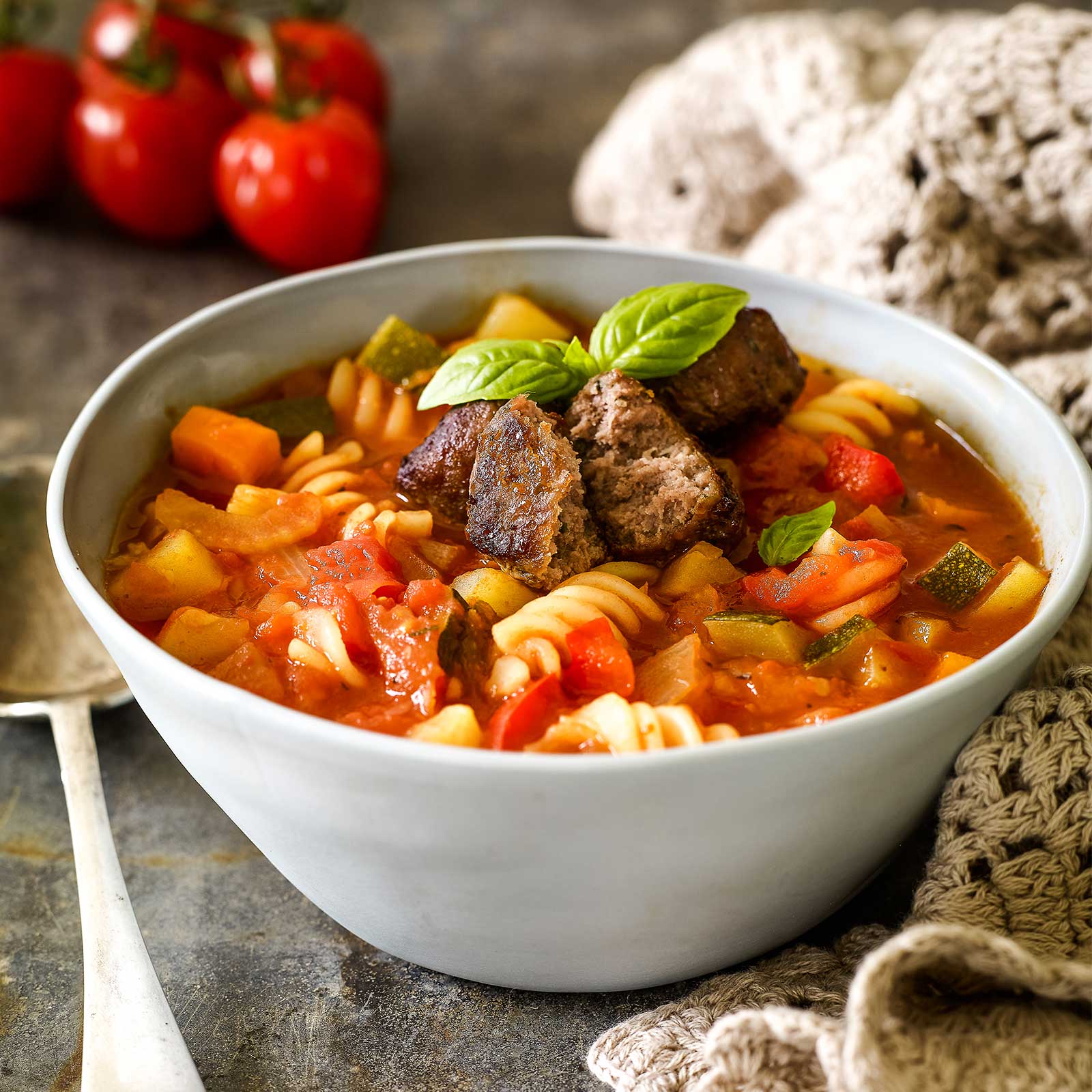 A light blue ceramic bowl is filled with gluten-free minestrone and topped with chopped meatballs and fresh basil leaves. Truss tomatoes sit behind the bowl.