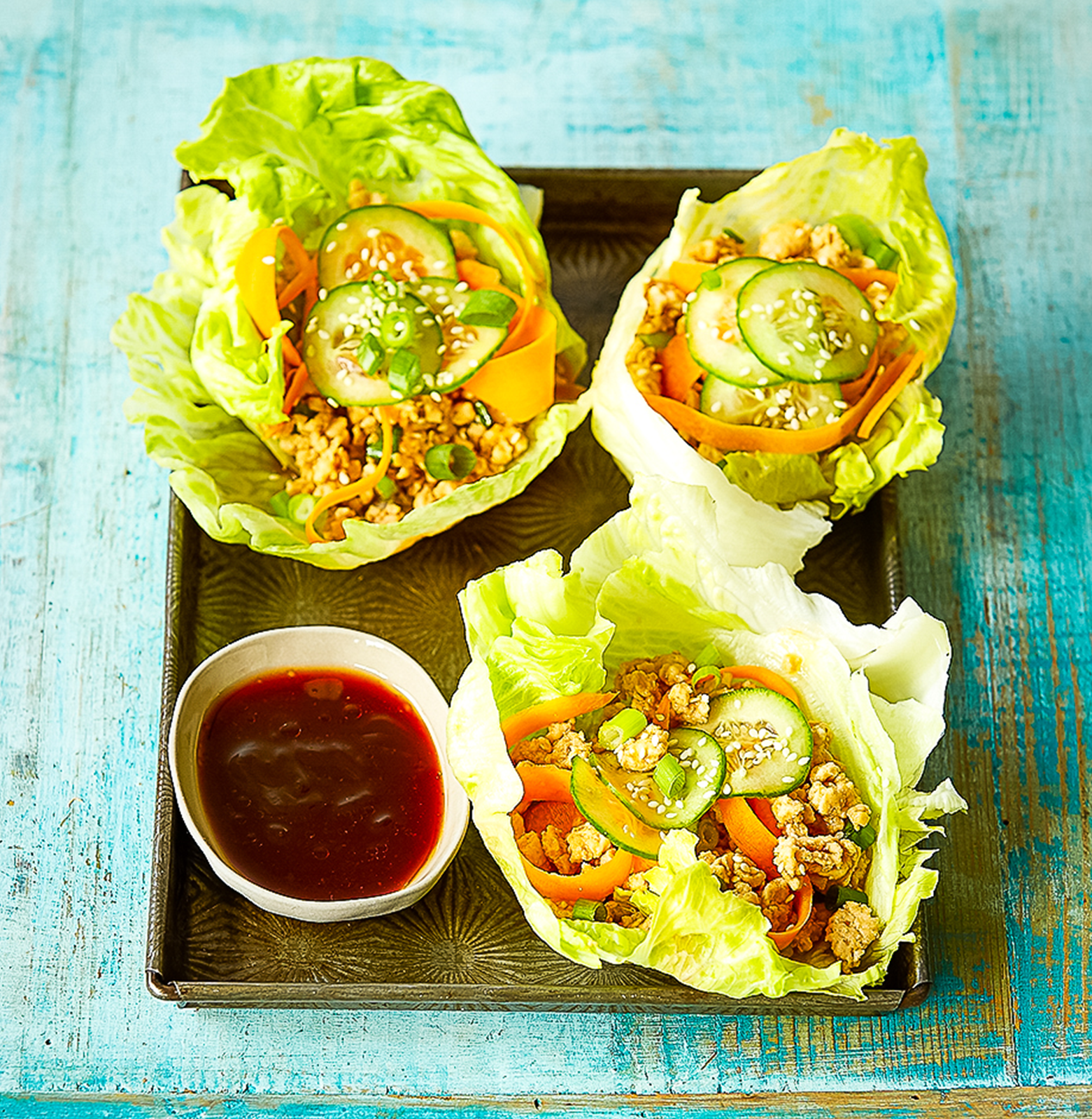 3 large iceberg lettuce leaves filled with spicy chicken mince filling arranged on a wooden board with a small dish of tamari and chilli sauce.