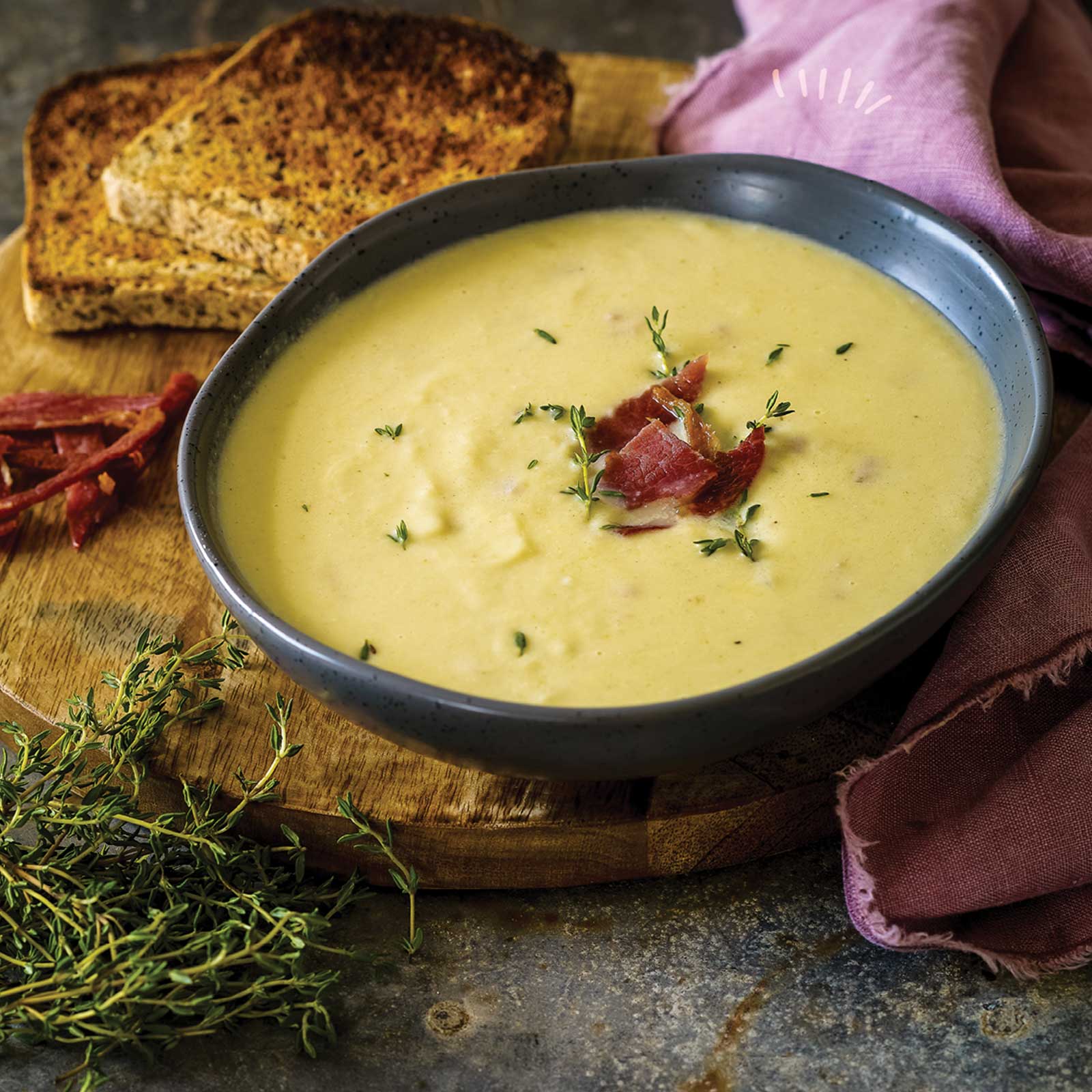 Dark blue bowl of creamy cauliflower and bacon soup sitting on a wooden board. Gluten-free toasts rests to the side. Fresh thyme leaves garnish the soup and the remaining herbs sit at the front of the image.
