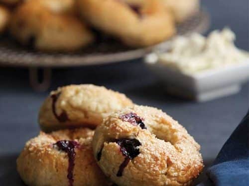 Gluten-free blueberry bagels are arranged on a round wire cooling tray at the back of the image. Just in front is a square container filled with creamy cheese. At the front of the image 3 gluten-free blueberry bagels are stacked ready to be eaten.