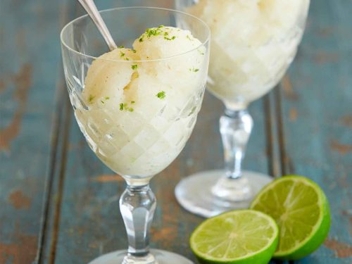Two crystal wine glasses are filled with scoops of lychee sorbet. Silver spoons are placed inside each glass ready to ear. A sliced lime sits in front of one class.