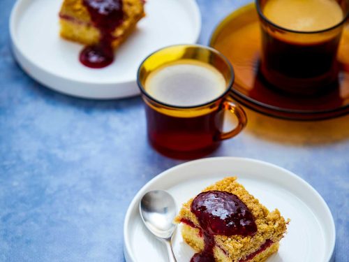 Dairy-free raspberry crumble squares sit on two round plates with an extra dollop of jam on top. Freshly made coffee is in between the plates in yellow glass espresso cups.