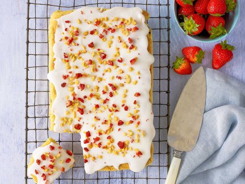 Dairy-free white chocolate brownie with white icing, crushed nuts and dried strawberries sits on a wire rack. A slice has been cut and is off to the side. Fresh strawberries are in a blue bowl at the back.