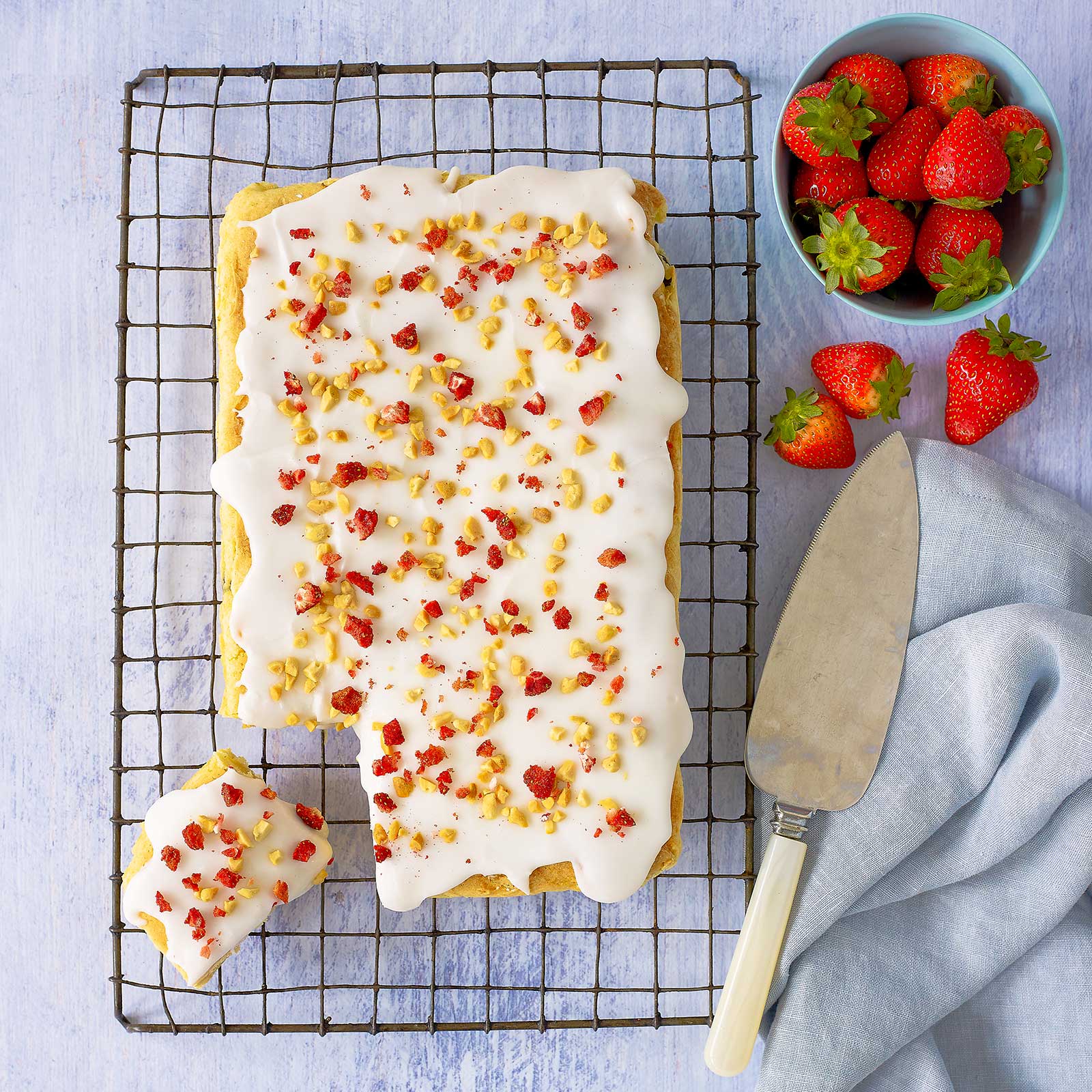 Dairy-free white chocolate brownie with white icing, crushed nuts and dried strawberries sits on a wire rack. A slice has been cut and is off to the side. Fresh strawberries are in a blue bowl at the back.