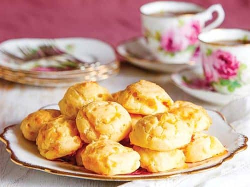 A morning tea setting with vintage plate, tea cups, saucers and embroidered napkins. On a large plate at the front is a tower of gluten-free Gougères de Bourgogne (gluten-free cheese puffs).