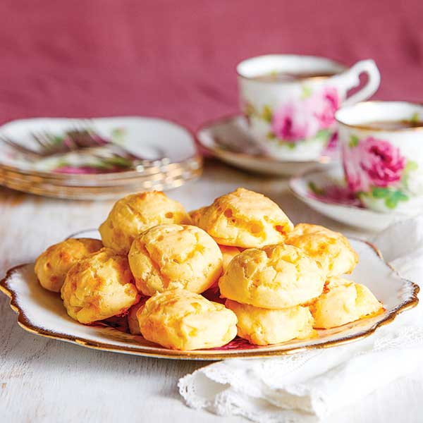 A morning tea setting with vintage plate, tea cups, saucers and embroidered napkins. On a large plate at the front is a tower of gluten-free Gougères de Bourgogne (gluten-free cheese puffs).