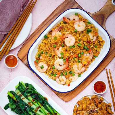 A large bowl of gluten-free fried rice with prawns is shot overhead. The dish rests on a wooden board. Chop sticks and tiny bowls of gluten-free chilli sauce are placed around the board. Steamed asian greens and a bowl of char siu chicken are just in the shot at the bottom.