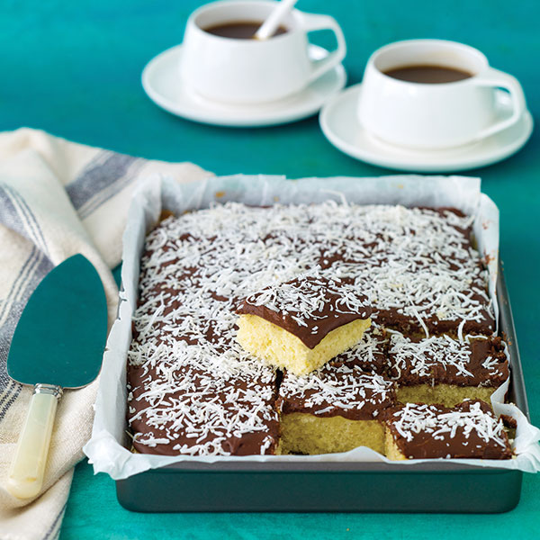 A lined baking tray rest on a aqua background. Inside the baking tray is an iced lamington slice (traybake). It has been cut into squares ready to serve and enjoy. In the background are two coffees in white cups resting on saucers.