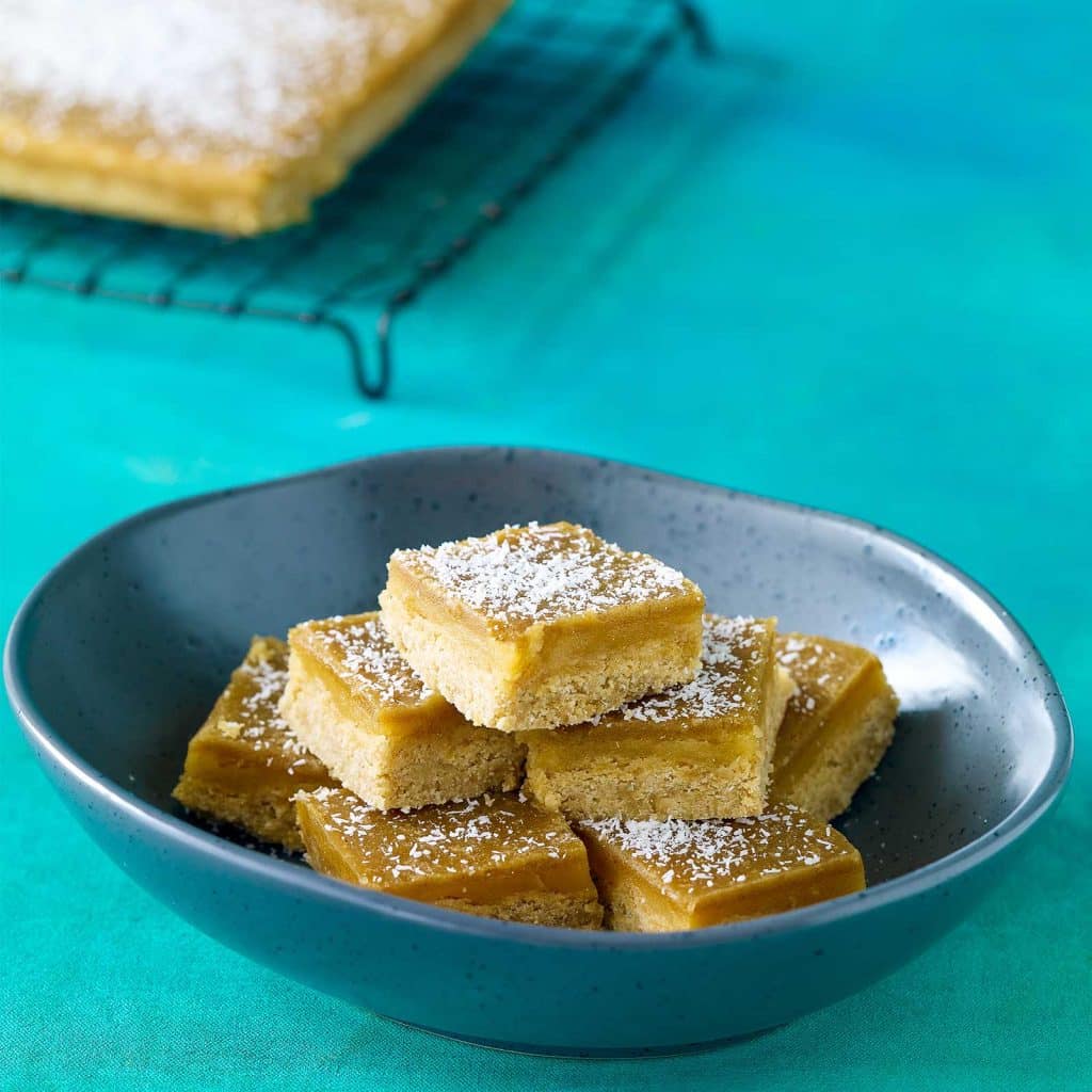 Squares of gluten-free ginger slice are arrange in a large bowl. The top of the slice has been sprinkled with coconut. The rest of the slice sits at the back on a wire cooling rack.