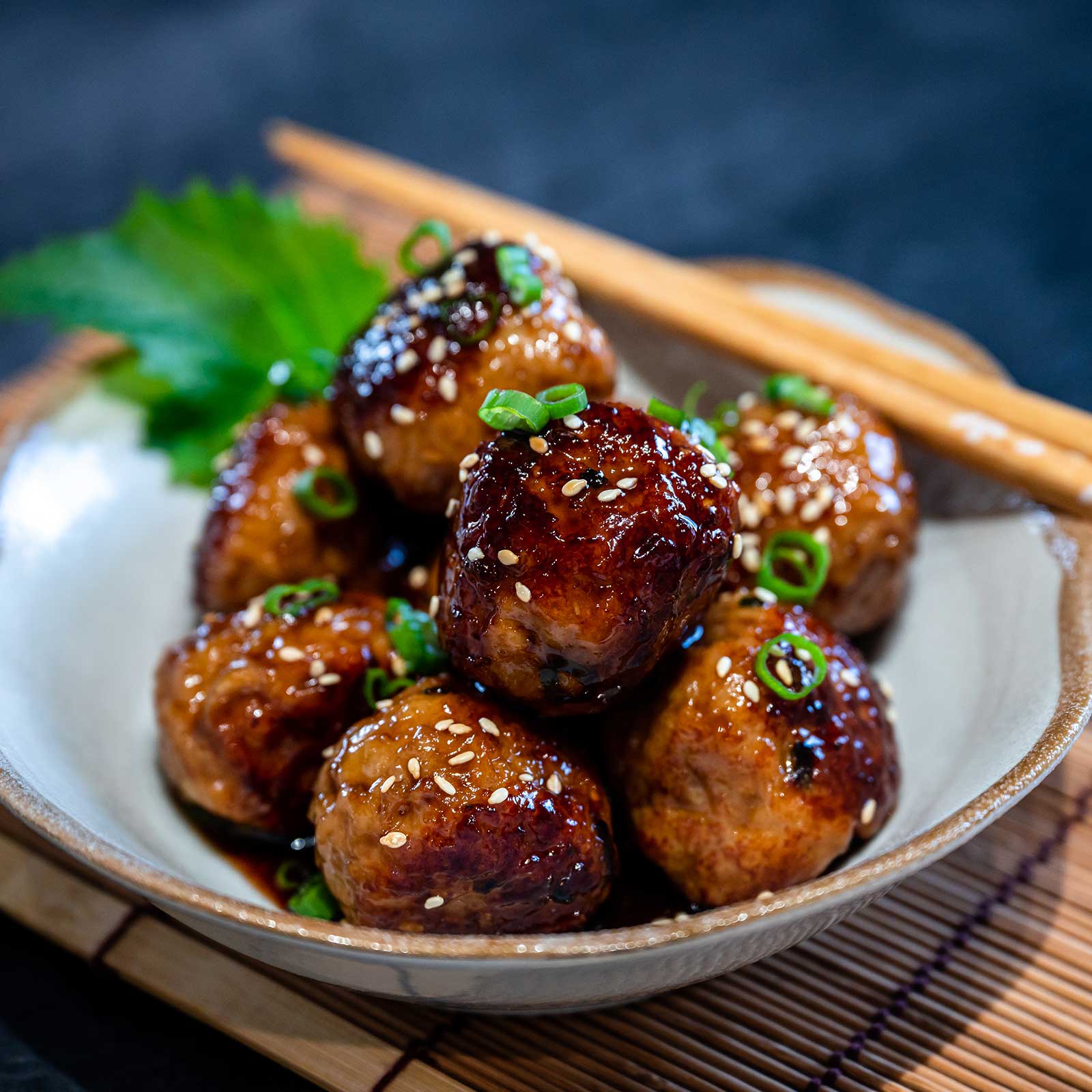 A ceramic bowl is filled with gluten-free japanese chicken meatballs. The light blue bowl is resting on a bamboo place mat and wooden chopsticks rest on the side of the bowl. The meatballs have been sprinkled with seseame seeds.