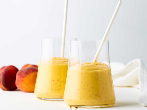 Two glass jars are half-full with spiced tropical smoothie. Both glasses hold a white paper straw. Two peaches sit at the back of the image behind the glasses.