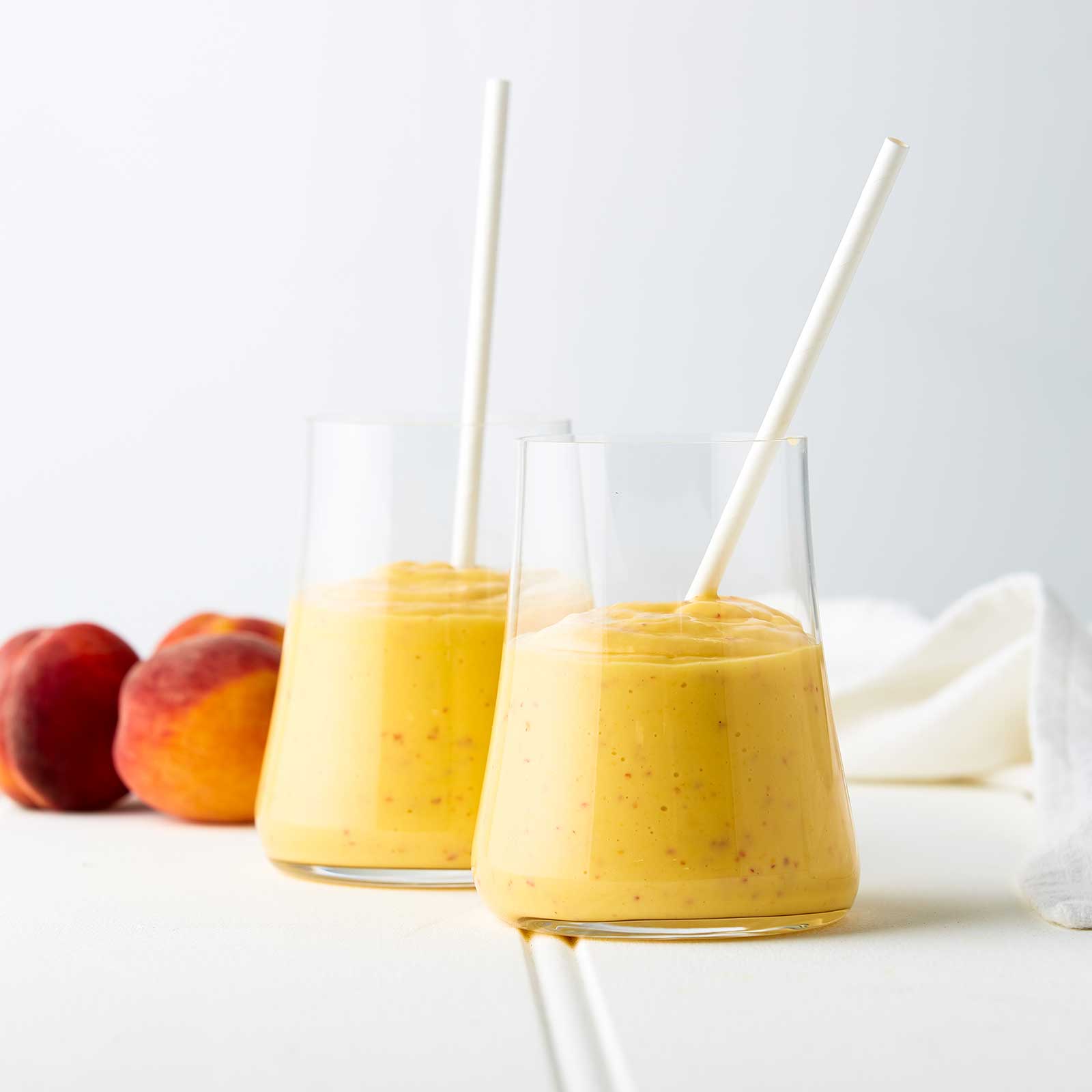 Two glass jars are half-full with spiced tropical smoothie. Both glasses hold a white paper straw. Two peaches sit at the back of the image behind the glasses.