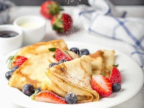 Gluten-free dairy-free crepes are folded and served on a white plate. They're topped with fresh berries and have been sprinkled with icing sugar. A breakfast setting is in the background.