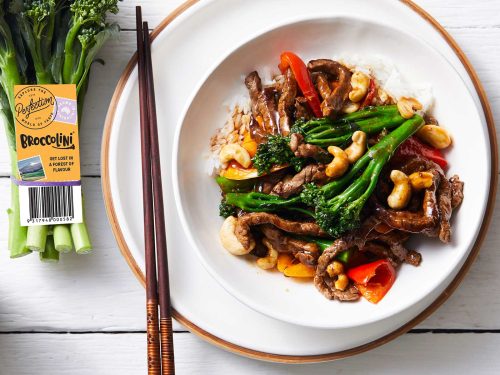 A white bowl with broccolini and teriyaki beef stir-fry sits on top of a flat white plate with wooden trim. Chop sticks rest on the left hand side of the flat plate. A bunch of bright green fresh broccolini rest in the top left corner.