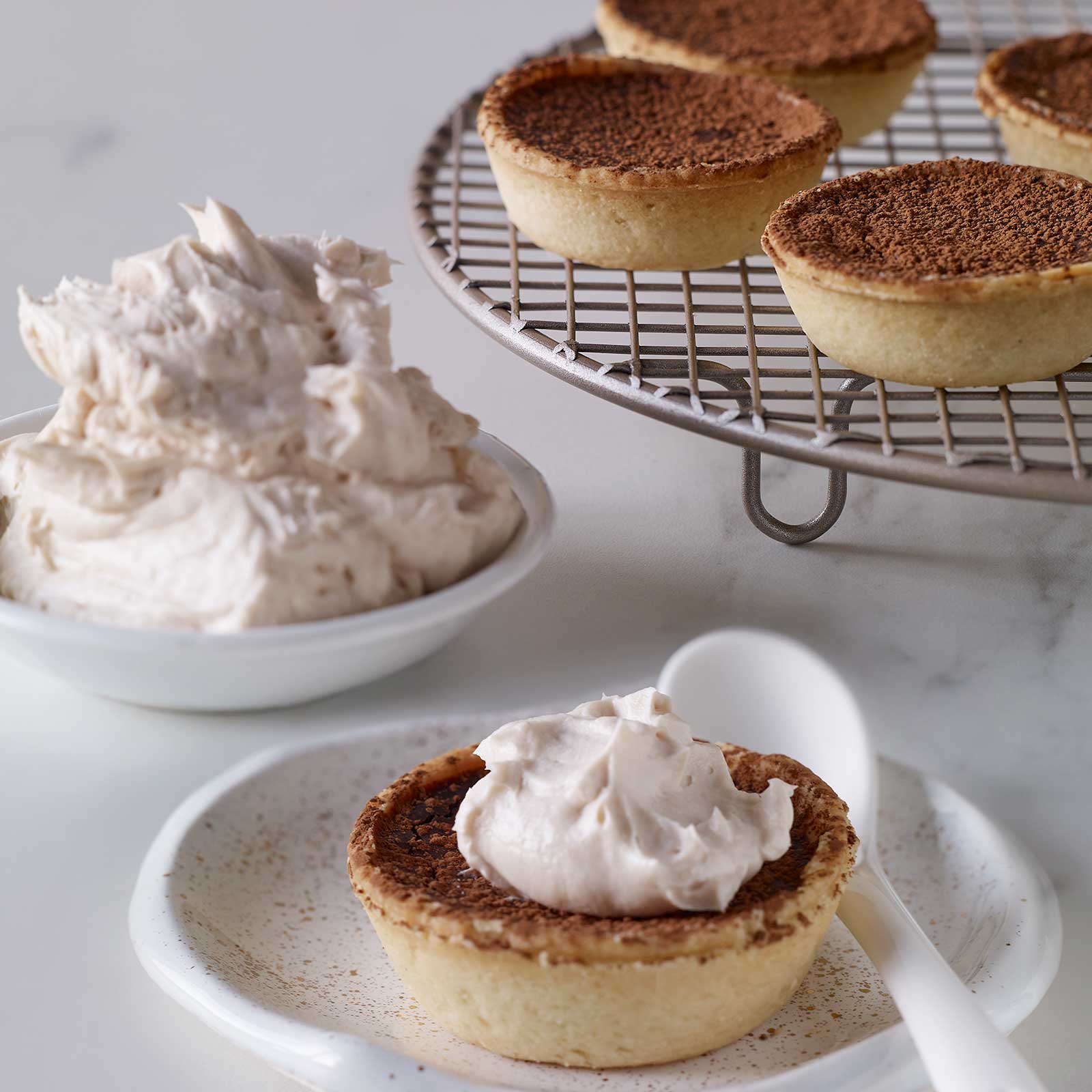 A small white plate with a gluten-free tart shell filled with gluten-free custard. A dollop of whipped cream sit on top with a sprinkle of cocoa. A wire rack sits at the back with more gluten-free chocolate tarts. A small bowl filled with whipped cream is next to the wire rack.