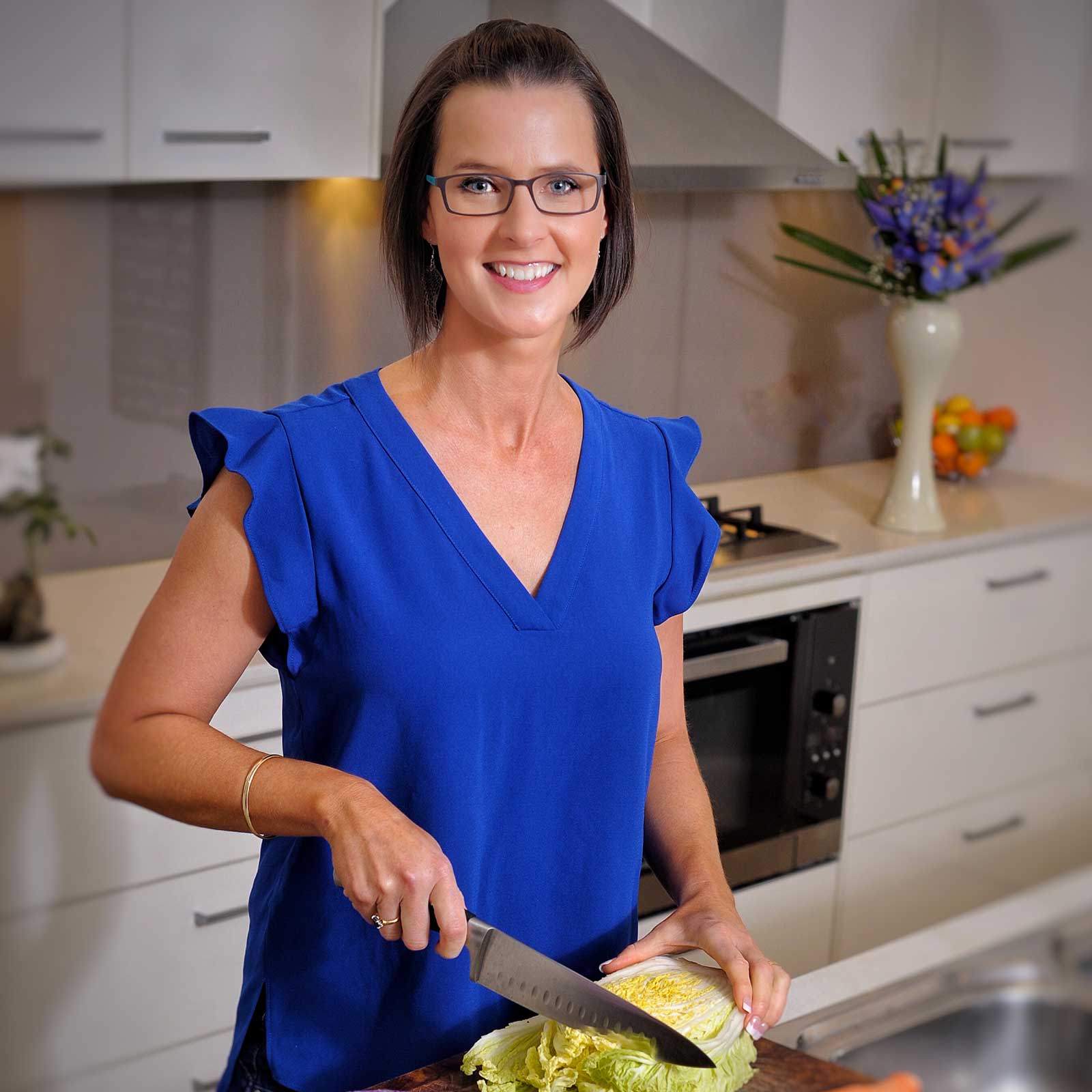 Allyson Browne is photographed in a kitchen slicing a cabbage. She wears a bright blue sleeveless shirt and reading glasses.