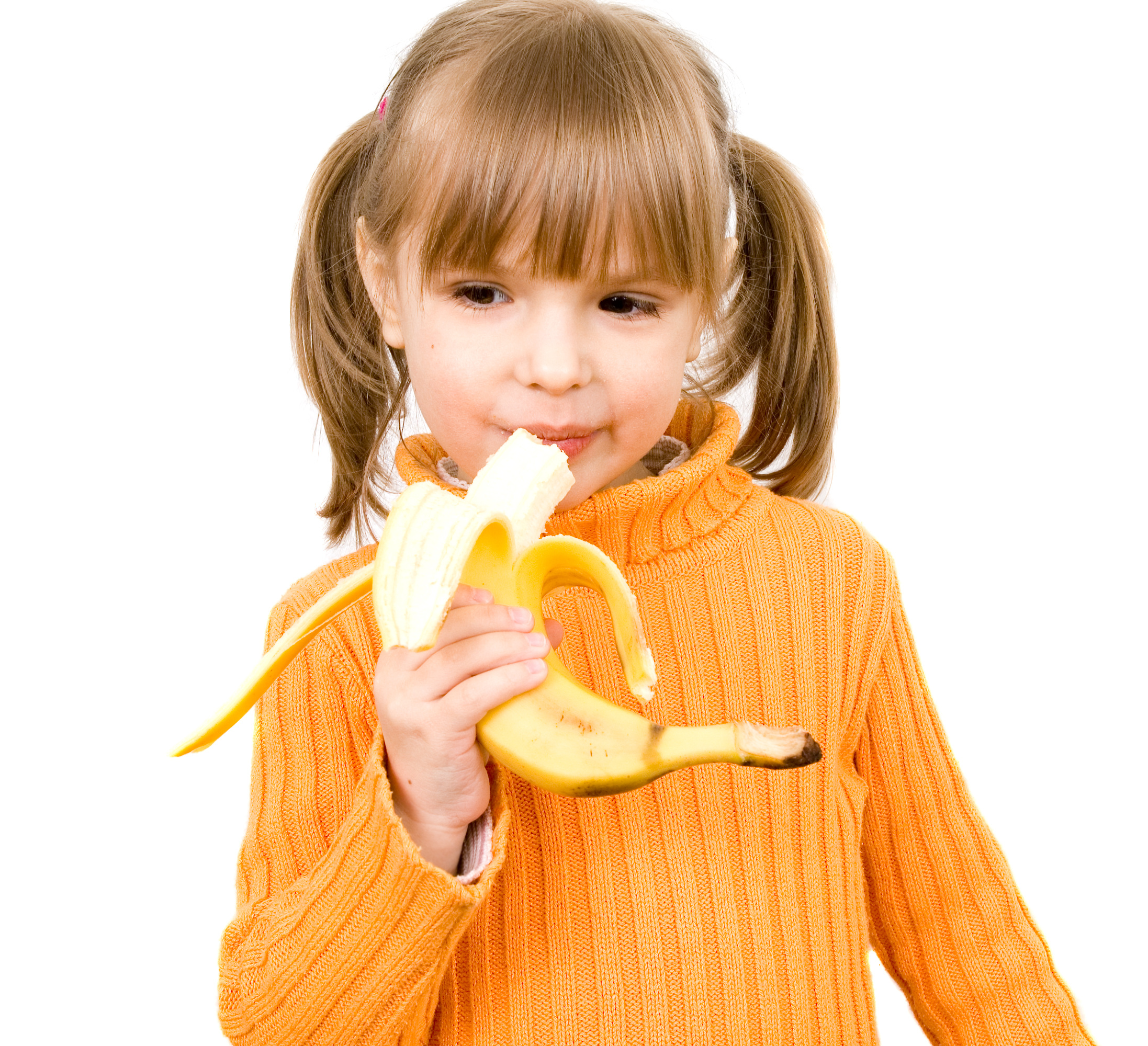 Young girl with pigtails in an orange jumper eats a banana.