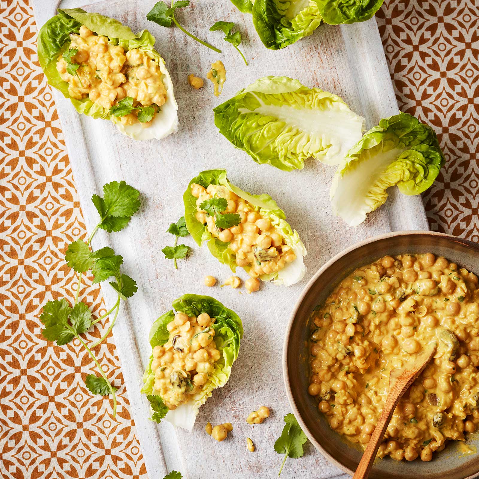 Coronation chickpea salad is in a saucepan with a wooden spoon. It sits on a white serving platter with lettuce leaves, some are filled with the chickpea mixture.