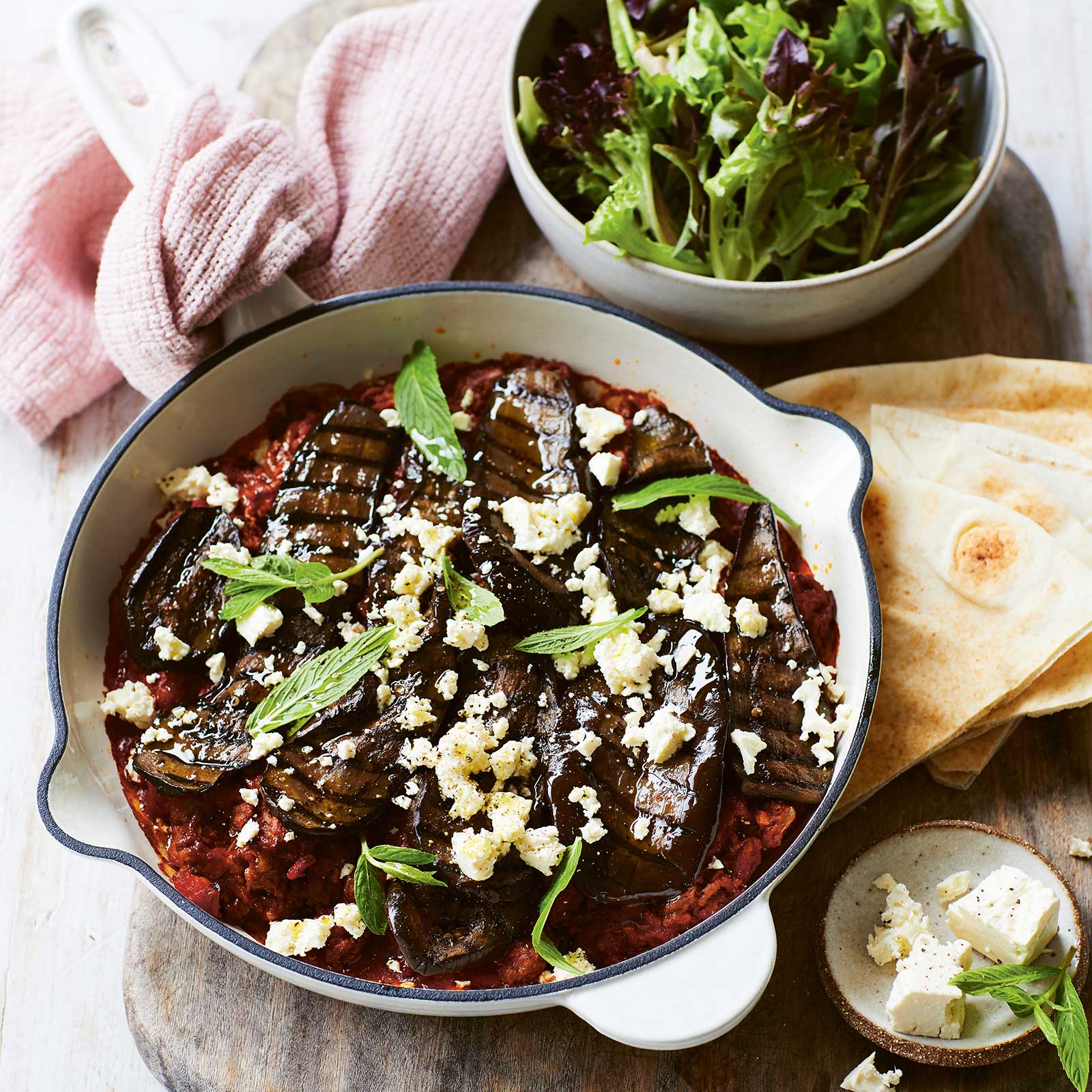A white cast-iron pan is filled with an easy moussaka. The top has been sprinkled with feta and fresh herbs. At the back is a small salad of leafy greens in a bowl and slices of gluten-free pita sit on the side.