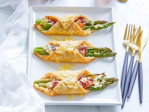 Three gluten-free prosciutto and asparagus bundles are lined up on a white serving platter. Black and gold cutlery rests next to the platter. At the back of the image a six-pack of gluten-free beer is ready to be enjoyed.