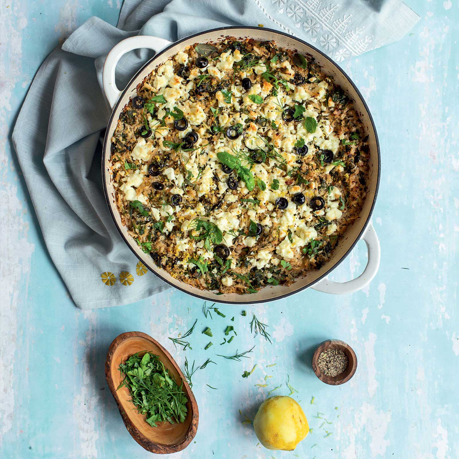 A recipe shot overhead. A white ceramic baking dish is filled with spanakorizo made from cauliflower instead of rice. Lots of fresh herbs, lemon and seasonings in the image.