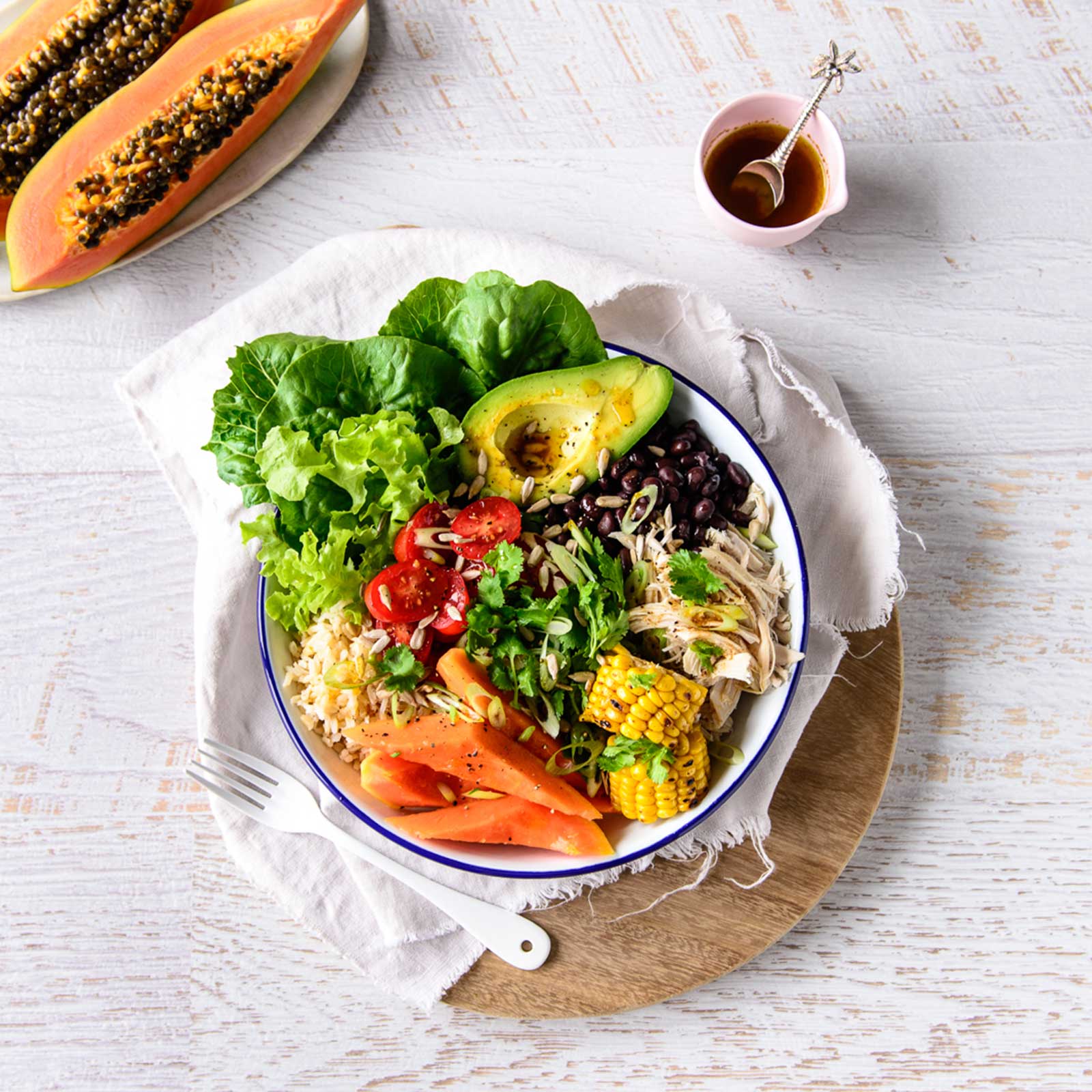 A white bowl with blue edge is shot over head. It rests on a round wooden board. A Mexican papaya buddha bowl is in the white bowl. A small dish with dressing rests to the side.