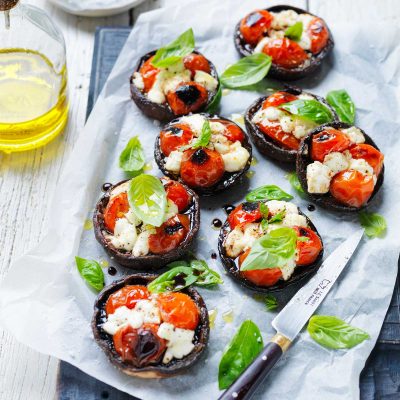 7 Portabella mushrooms are arranged on a board lined with baking paper. The mushrooms have been stuffed with cheese, tomatoes and basil and baked. The mushrooms are garnished with fresh basil leaves. A knife and a bottle of oil are positioned at the side.