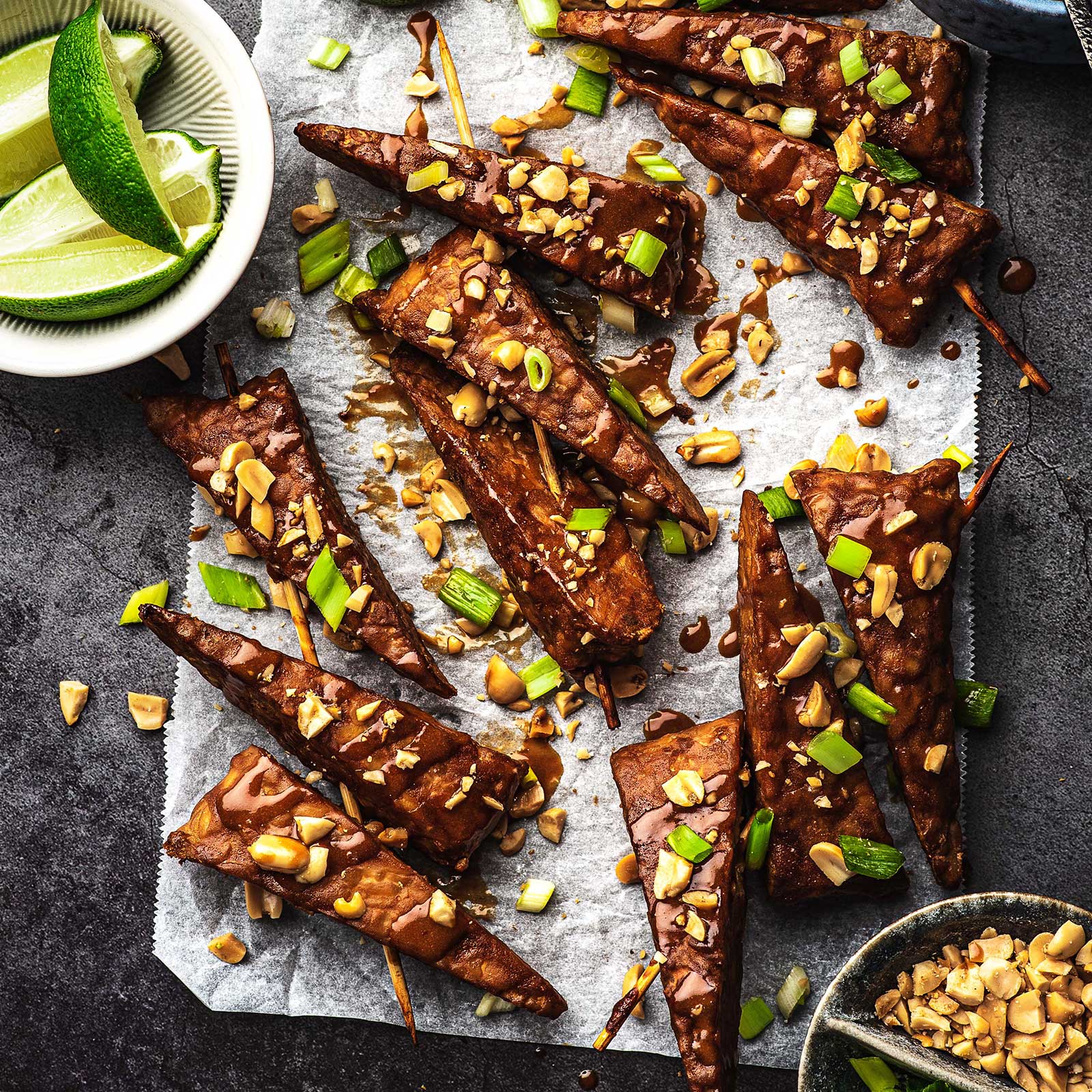Wedges of smokey tempeh peanut satay are arranged on a piece of baking paper. The wedges have been garnished with crushed peanuts and spices of spring onion. A bowl of lime wedges sit at the side.