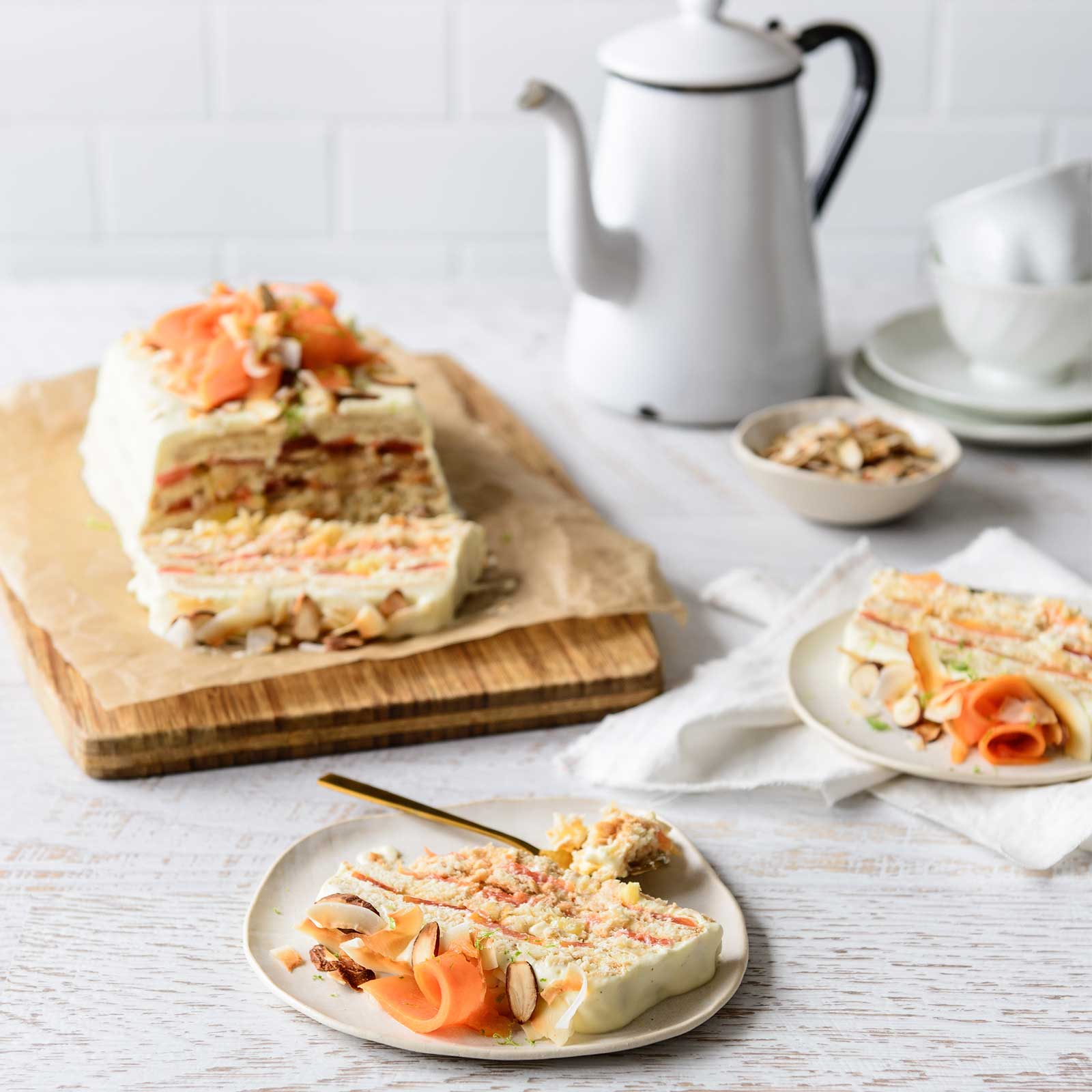 Gluten-free tropical papaya refrigerator cake sits on a wooden board at the back of the image. Two slices are sitting on places at the front of the board. At the back is a vintage enamel coffee pot awithnd cups and sauces.