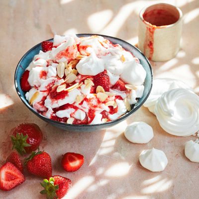 British summertime dessert Vegan Eton Mess is served in a bowl and topped with fresh strawberries and toasted coconut flakes. Fresh strawberries and extra vegan meringues sit around the bowl.
