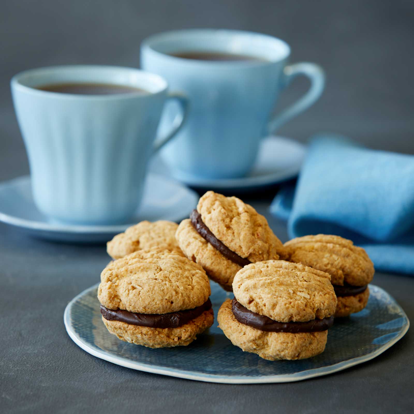 Five vegan and gluten-free kingston biscuits are arranged on a round blue plate. In the background two blue mugs sitting on saucers are filled with black tea.