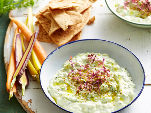 Two bowls of avocado tzatziki rest on a large wooden board. Dutch carrots and gluten-free pita bread rest on the side.