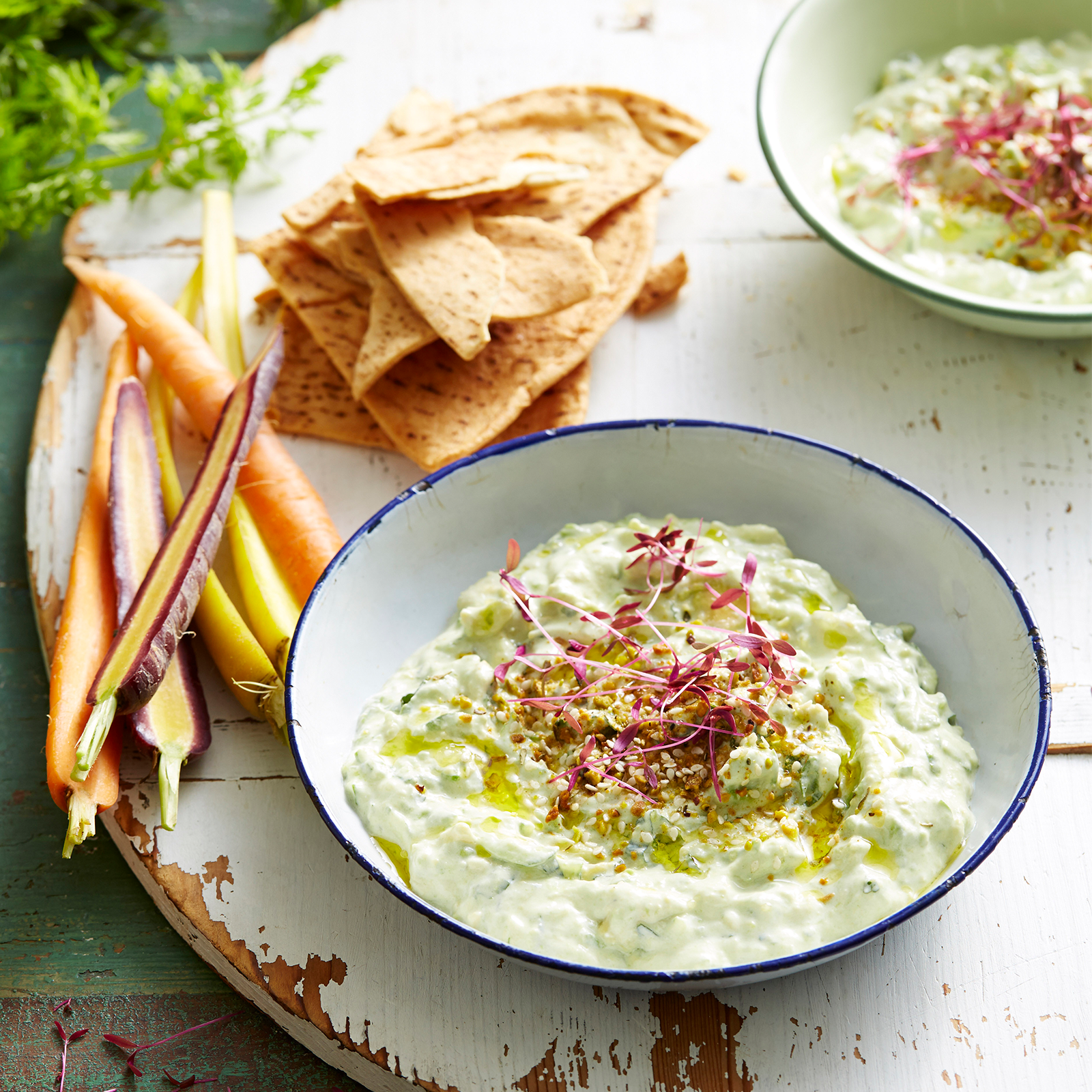 Two bowls of avocado tzatziki rest on a large wooden board. Dutch carrots and gluten-free pita bread rest on the side.