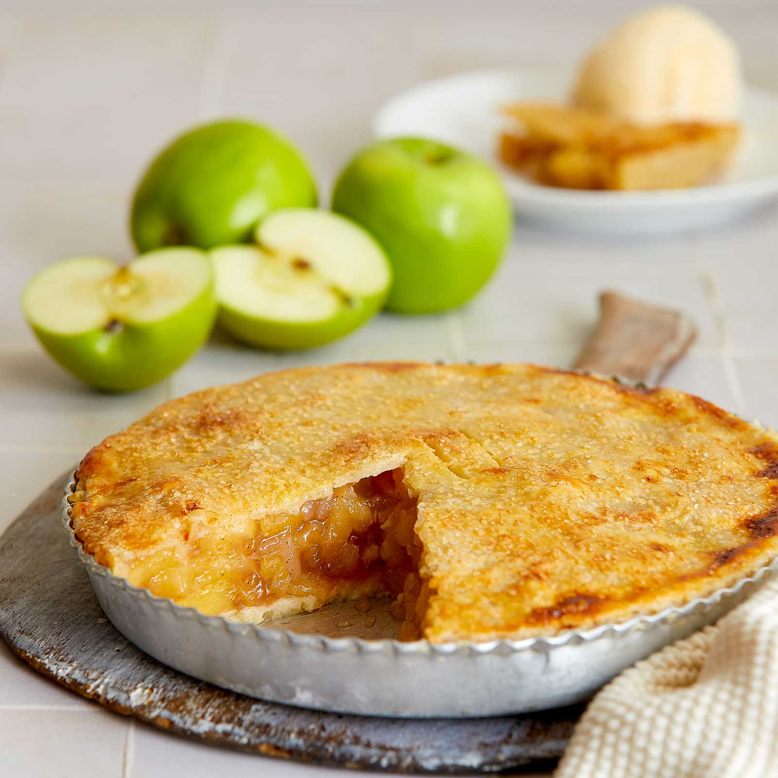 A silver pie dish rests on a wooden tray. A gluten-free apple cider pie has been baked in the pie dish. A slice has been removed and is plated at the back with a scoop of ice-cream. Some Granny Smith Apple rest between the pie and served slice.